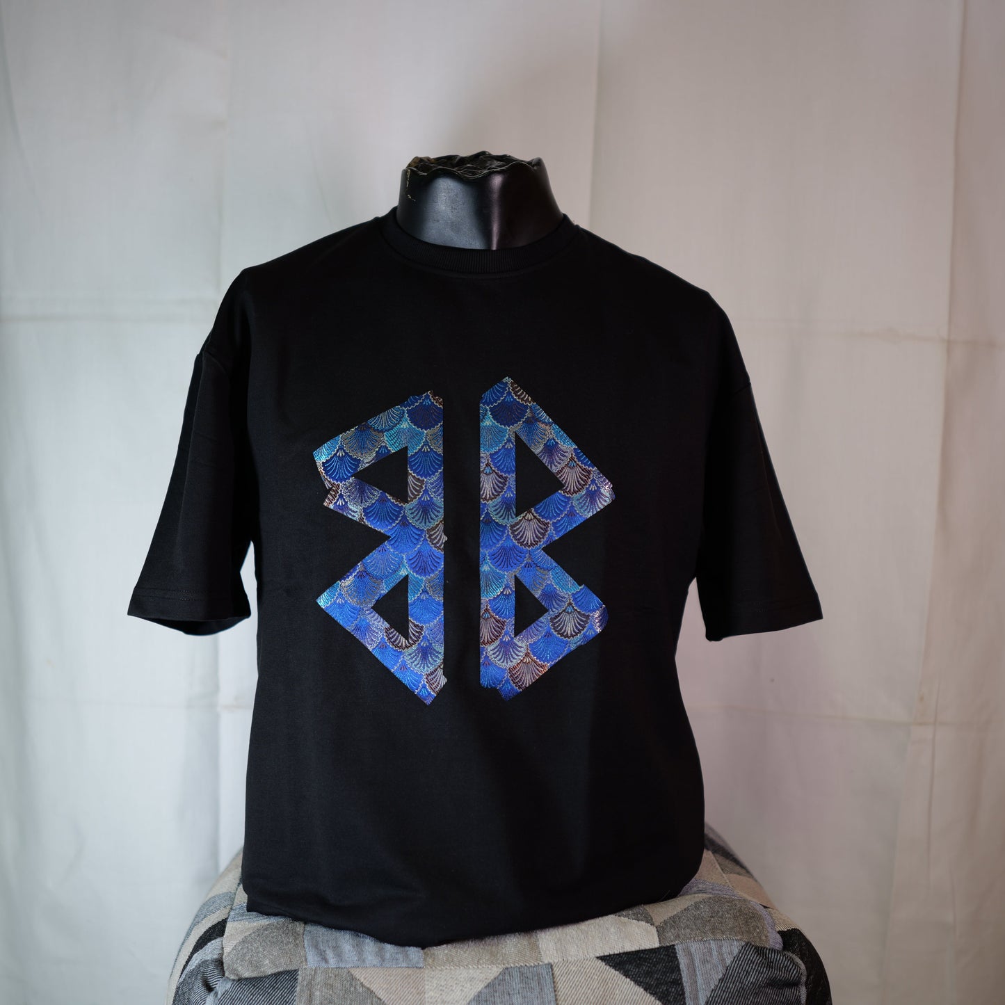 Black t-shirt with blue geometric design on a mannequin against a white curtain.