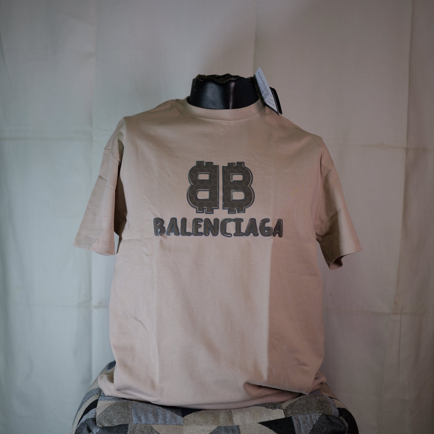 Beige t-shirt with Balenciaga logo on a mannequin against a white curtain.